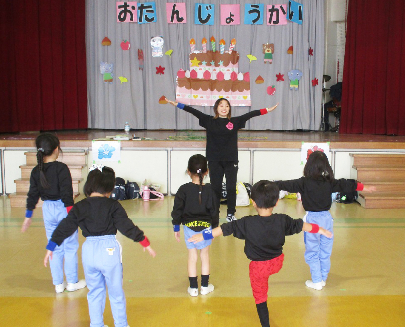 ジャージ姿の先生と子どもたちが両手を広げています。