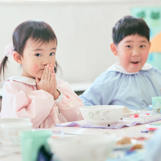 手を合わせてカメラの方を見る女の子と、その後ろで男の子が微笑んでいます。