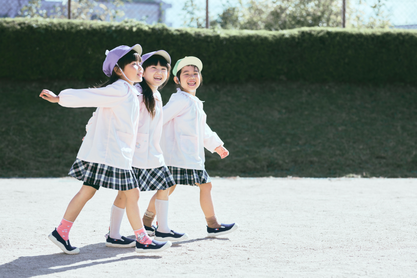 3人の女の子が手を繋いで楽しそうな表情で園庭を歩いています。